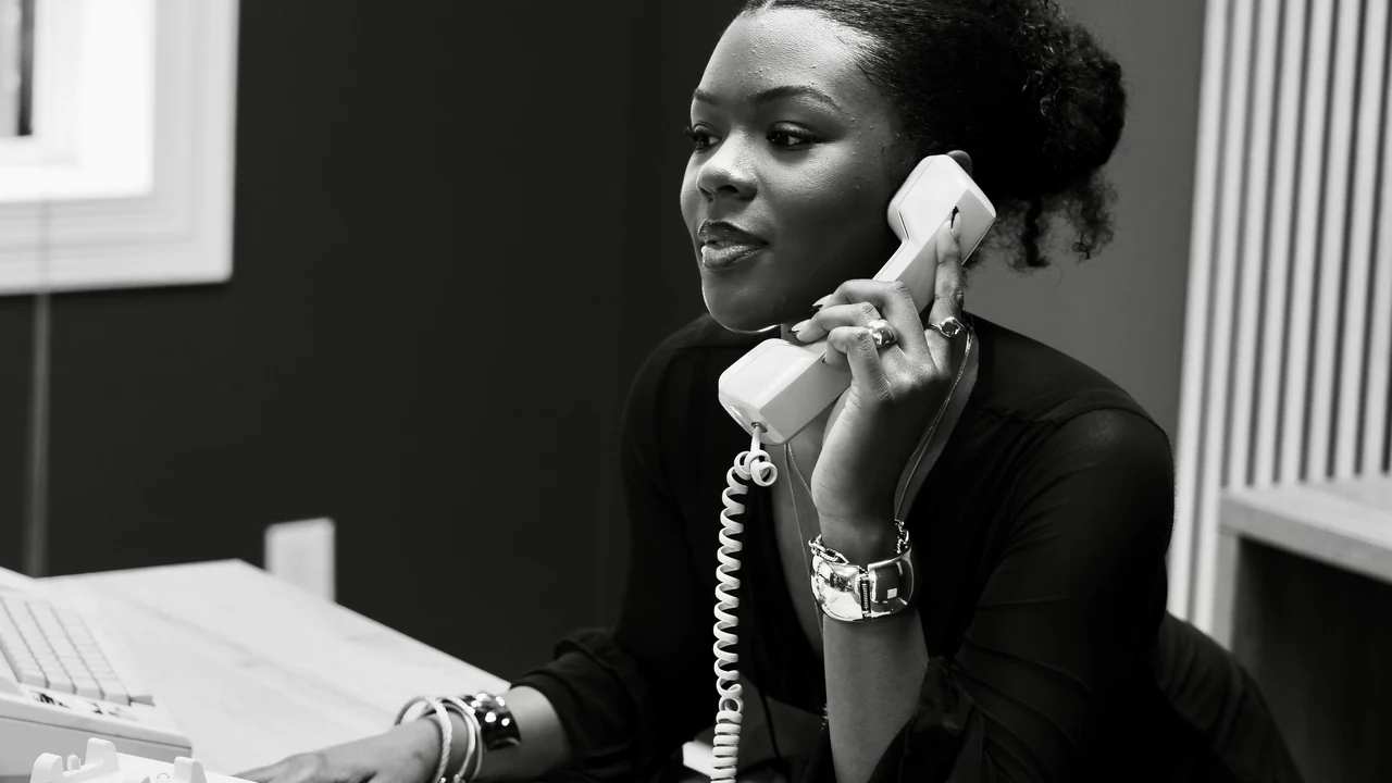 Woman talking on a vintage telephone at a desk.